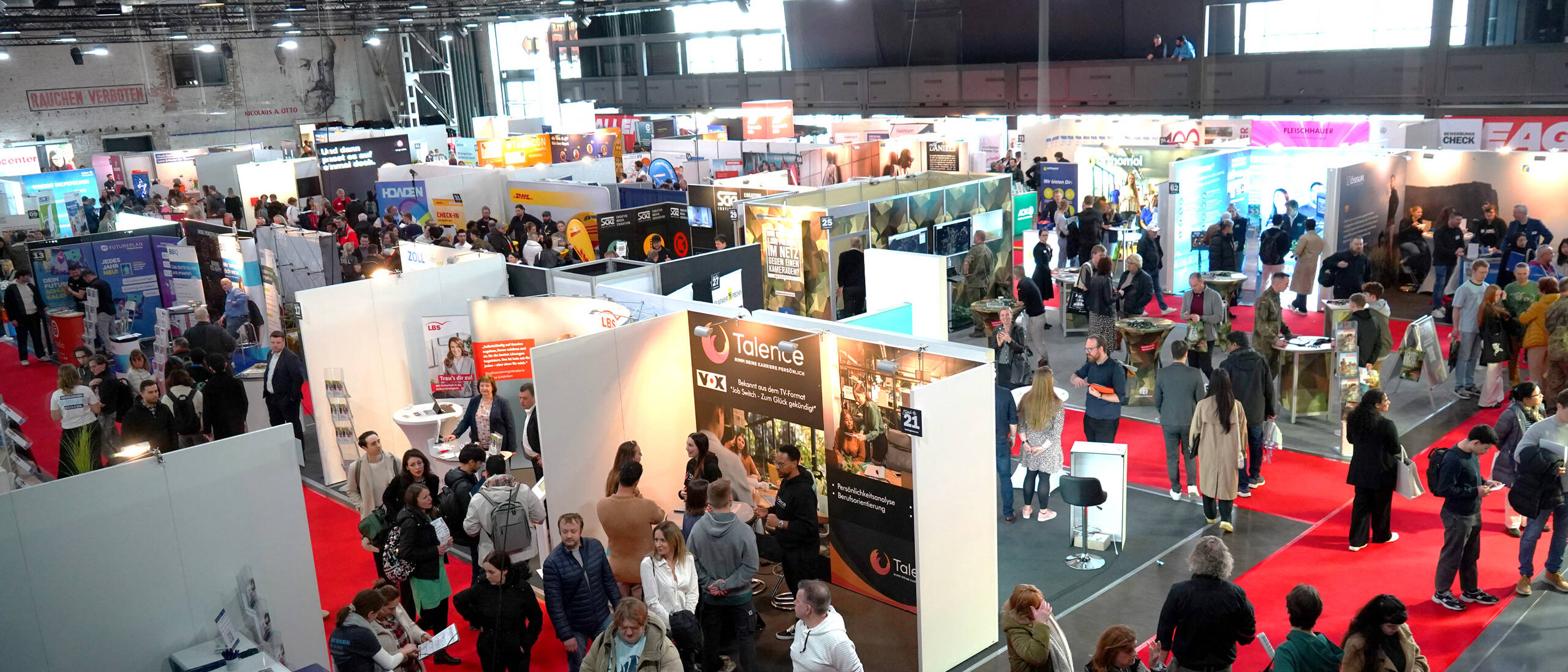 Messehalle mit vielen Ausstellungsständen und Besuchern, rote Gänge und Menschen, die sich an Messeständen informieren.