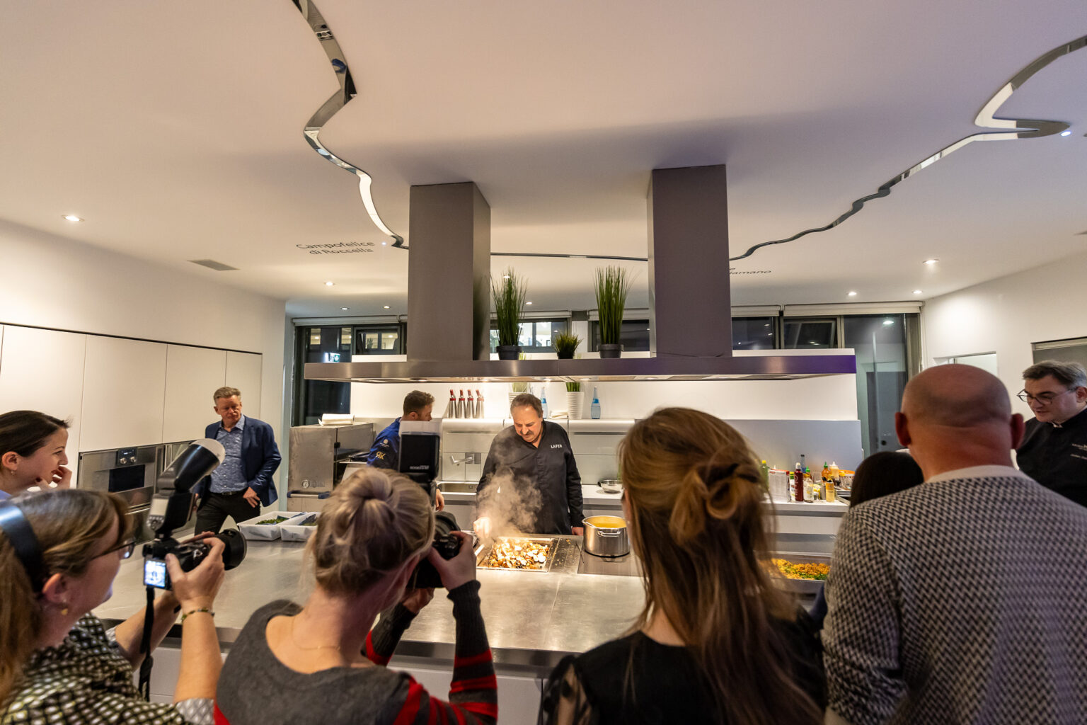 Koch demonstriert Gerichte an einer Kücheninsel, umringt von Fotografen und Zuschauern.