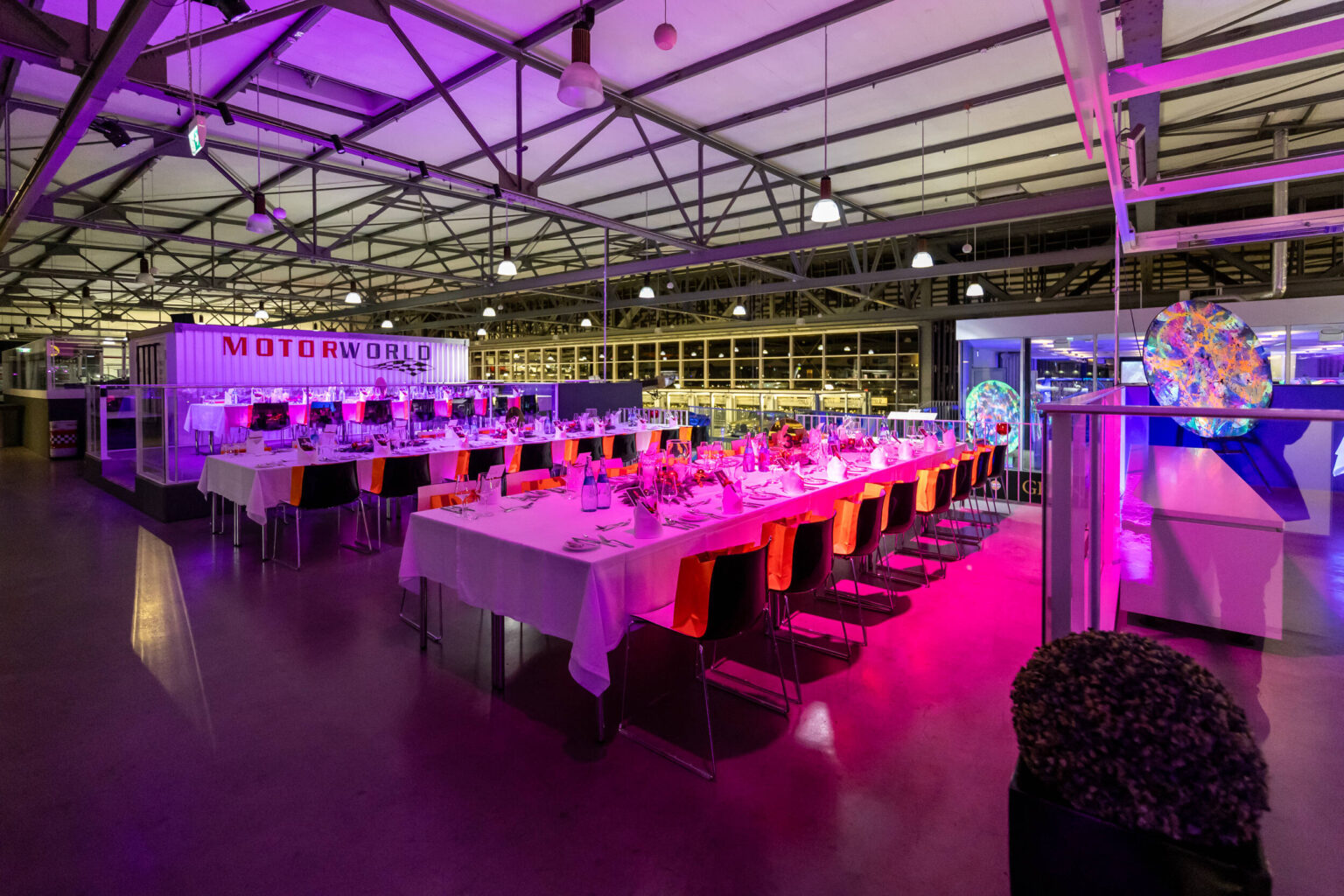 Große Eventhalle mit lang gedeckten Tischen, buntem Pink- und Violettlicht und sichtbarer Stahlträgerdecke.