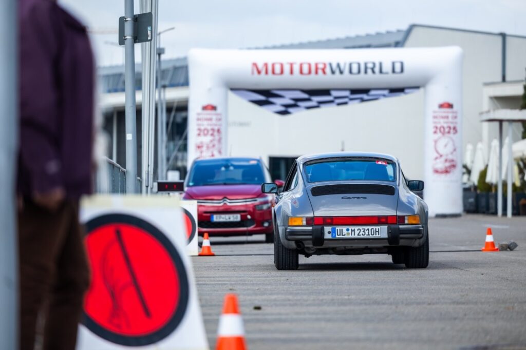 Silberner Porsche 911 Carrera von hinten fährt durch Absperrung mit Motorworld-Startbogen im Hintergrund.