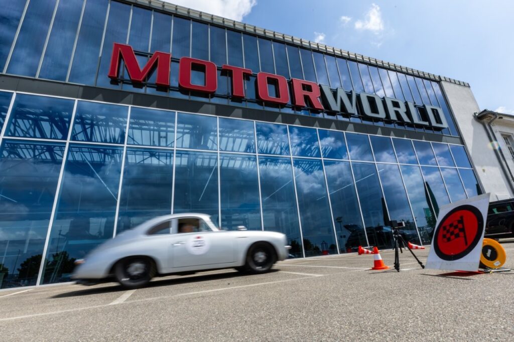 Silbernes Oldtimer-Coupé fährt vor dem gläsernen Gebäude mit großem Schriftzug „MOTORWORLD“.