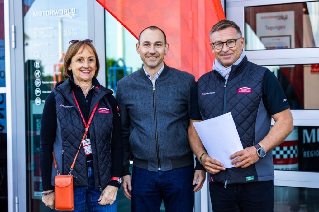 Drei lächelnde Erwachsene in Outdoorweste vor einem Gebäudeeingang, die rechte Person hält Papiere.