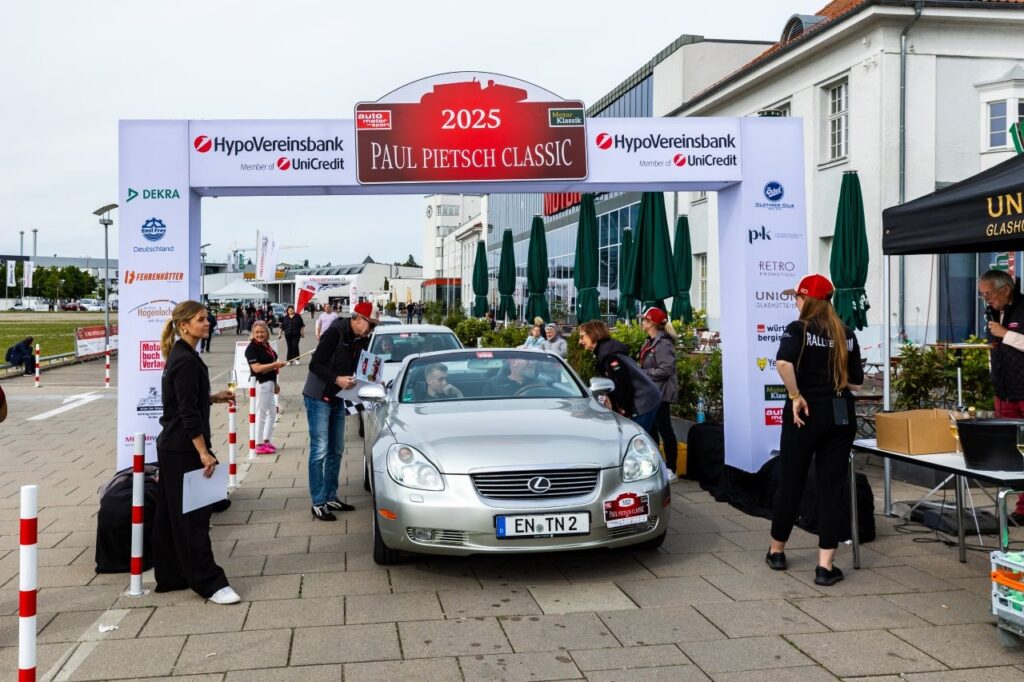 Silberner Lexus fährt unter Startbogen "Paul Pietsch Classic 2025", Veranstalter und Helfer kontrollieren Teilnehmer.