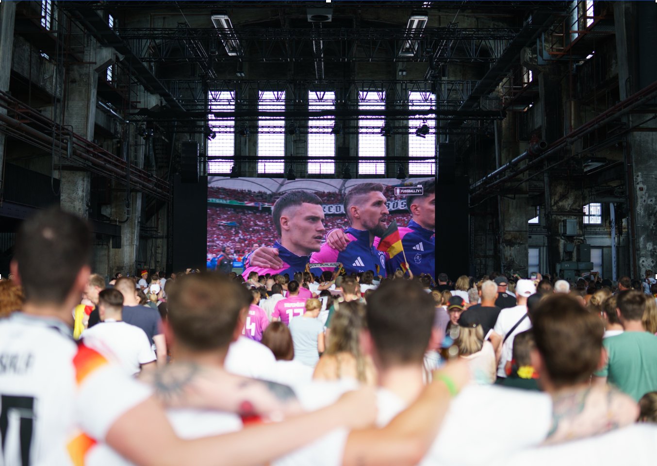 Menschenmenge schaut in einer Industriehalle ein Fußballspiel auf einer großen Leinwand