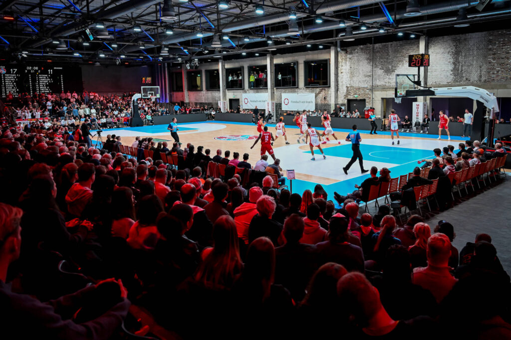 Basketballspiel in einer vollen Halle mit jubelndem Publikum und laufender Aktion auf dem Spielfeld