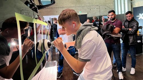 Junger Mann in weißem T-Shirt und Ohrhörern poliert die Fahrzeugseite eines schwarzen Autos mit einem Poliergerät, während zwei Männer im Hintergrund zusehen und einer davon ein Smartphone zum Filmen hält.