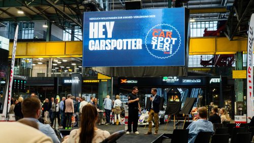 Veranstaltungsraum mit Publikum und zwei Sprechern auf einer Bühne unter einem großen Bildschirm mit dem Text "Hey Carspotter" und dem Logo des Carspotter Awards 2025, im Hintergrund Industriehalle mit gelbem Stahlträger und verschiedenen Firmenschildern.