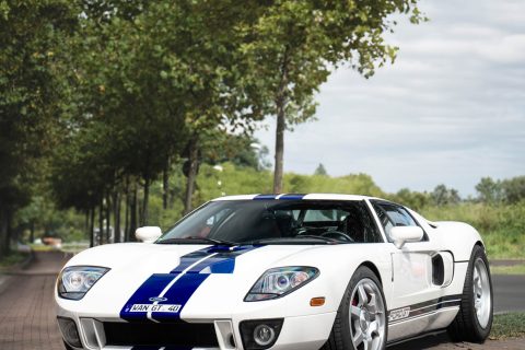 Weißer Ford GT Sportwagen mit blauen Rennstreifen, geparkt auf gepflastertem Weg neben Baumreihen unter bewölktem Himmel.