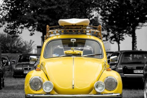 Vorderansicht eines gelben Volkswagen Käfer mit Dachgepäckträger und Surfbrett, geparkt auf einer Wiese, mit schwarz-weiß gehaltenem Hintergrund.