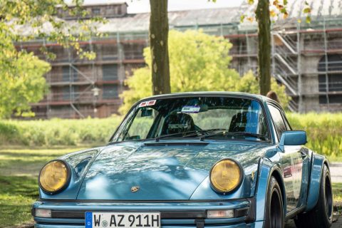 Blauer Porsche 911 mit gelben Scheinwerfern und breiten Radläufen, geparkt auf einem Straßenrand mit Bäumen und einem Gebäude im Hintergrund.