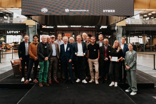 Gruppenfoto von Preisträgern und Gästen in einer Ausstellungshalle mit Automobilmarken im Hintergrund