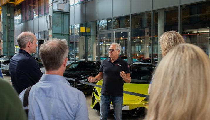 Man in black polo explains in front of a group and a yellow sports car in a hall.
