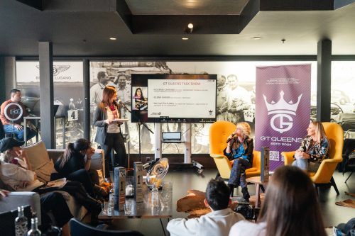 Frauen-Podiumsdiskussion bei der GT Queens Talk Show mit einer Moderatorin stehend neben einem Bildschirm, zwei Frauen in gelben Sesseln mit Mikrofonen, Publikum im Vordergrund und großem GT Queens-Banner im Hintergrund.