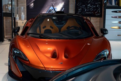Roter McLaren-Sportwagen mit orangefarbenen Sitzen in einem Showroom mit Steinwand und Dekoration im Hintergrund, teilweise von einem schwarzen Auto im Vordergrund verdeckt.