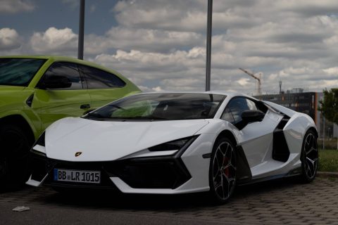 Weißer Lamborghini-Sportwagen, geparkt auf einem Pflaster neben einem grünen Auto unter einem bewölkten Himmel mit Laternen und einem Gebäude im Hintergrund.