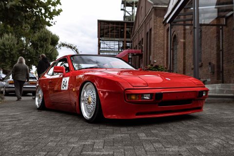 Roter Porsche 944 Sportwagen mit Rennnummer 84, abgestellt auf einem gepflasterten Platz vor einem Backsteingebäude mit Industriearchitektur im Hintergrund, links Bäume und zwei Menschen zu sehen