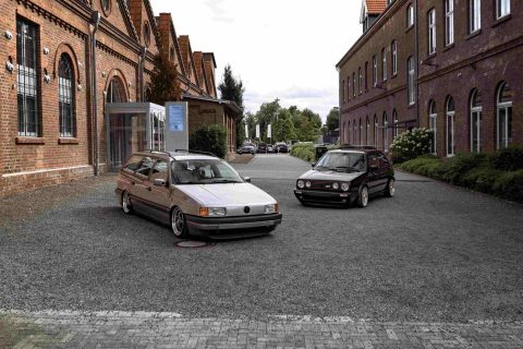 Zwei ältere, tiefgelegte Volkswagen-Kombis, geparkt auf einem geschotterten Hof zwischen zwei Reihen roter Backsteinbauten mit spitzen Dächern und runden Fenstern, bei bewölktem Himmel.