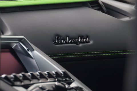 Nahaufnahme des Innenraums eines Lamborghini-Sportwagens mit Fokus auf das Armaturenbrett und die Mittelkonsole, schwarzes Leder mit grüner Ziernaht und Lamborghini-Logo sichtbar