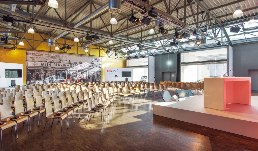 Heller, moderner Konferenzsaal mit zahlreichen in Reihen aufgestellten hellen Holzstühlen, einer erhöhten Bühne mit rosafarbenem Pult rechts, sichtbarer Stahlträger- und Lüftungsdecke sowie einer großen historischen Wandgrafik mit der Aufschrift MEIN BENZIN – GASOLIN links.