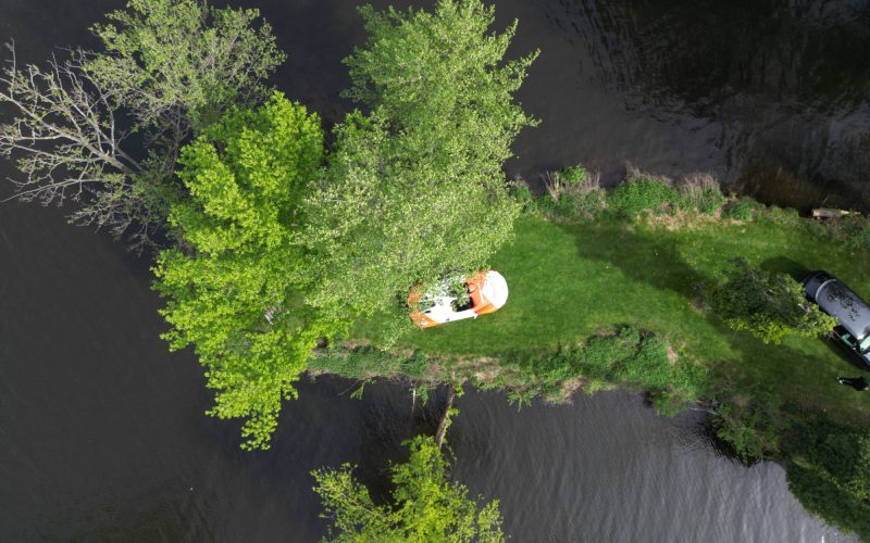 Aerial view of a narrow, green path between water surfaces with a white and orange bicycle and a parked car on the grass, surrounded by trees with green leaves.