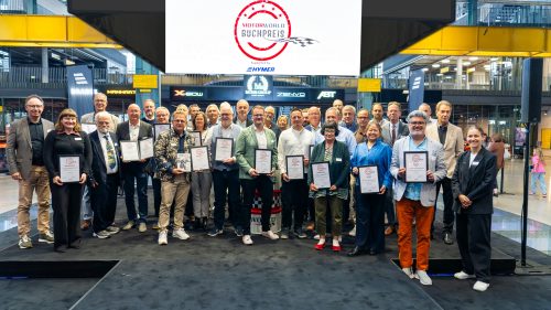 Große Gruppe von Menschen, viele mit Urkunden in der Hand, posieren auf einem schwarzen Teppich in einer modernen Ausstellungshalle mit gelben Balken und Schildern im Hintergrund, darüber ein Bildschirm mit dem Logo 'Motorworld Buchpreis'.