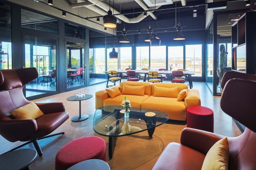 Motorworld_Mallorca_Drivers_Lounge_023 Moderner Besprechungsraum mit gelbem Sofa, roten Sesseln, bunten Stühlen und großem Tisch vor Panorama-Fenstern, die eine Landschaft zeigen.