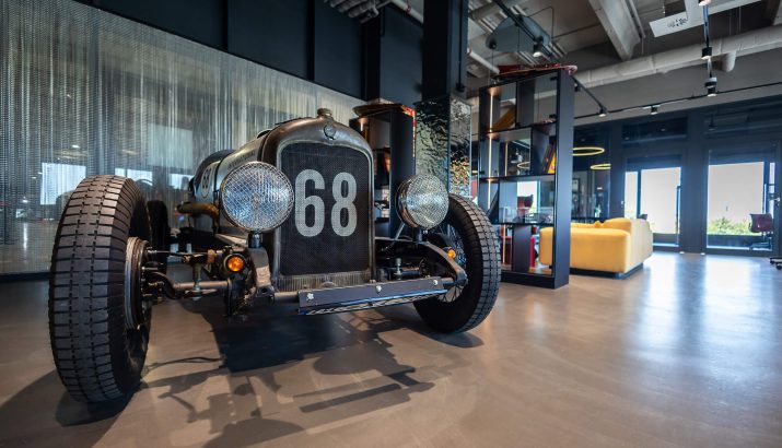 Nahaufnahme eines alten Rennwagens mit der Nummer 68 auf dem Kühlergrill, ausgestellt in einem modernen Innenraum mit großen Fenstern, Regalen und einer gelben Couch im Hintergrund.