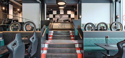 Rennsport‑themenlokal mit Treppe zum Schriftzug ZIEL, Rennsitze, ausgestellte Reifen und schwarz‑weiß kariertes Dekor.