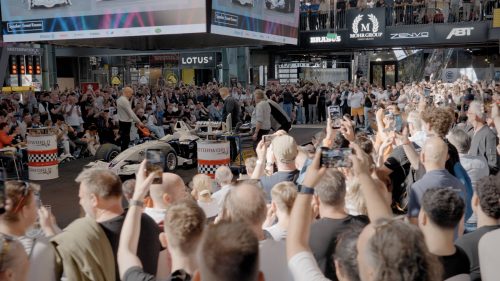 Große Menschenmenge filmt und fotografiert ein Rennwagen-Event in einer Halle mit Bühne und Werbebannern.