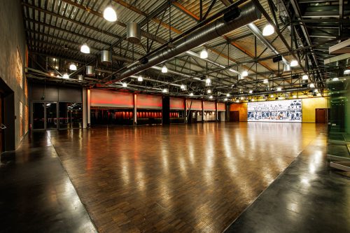Großer leerer Industrie-Eventsaal mit glänzendem Parkettboden, sichtbaren Metallträgern und Lüftungsrohren an der Decke, zahlreichen Hängeleuchten, roter Akzentbeleuchtung an der Rückwand und einer großen leuchtenden Wandgrafik.