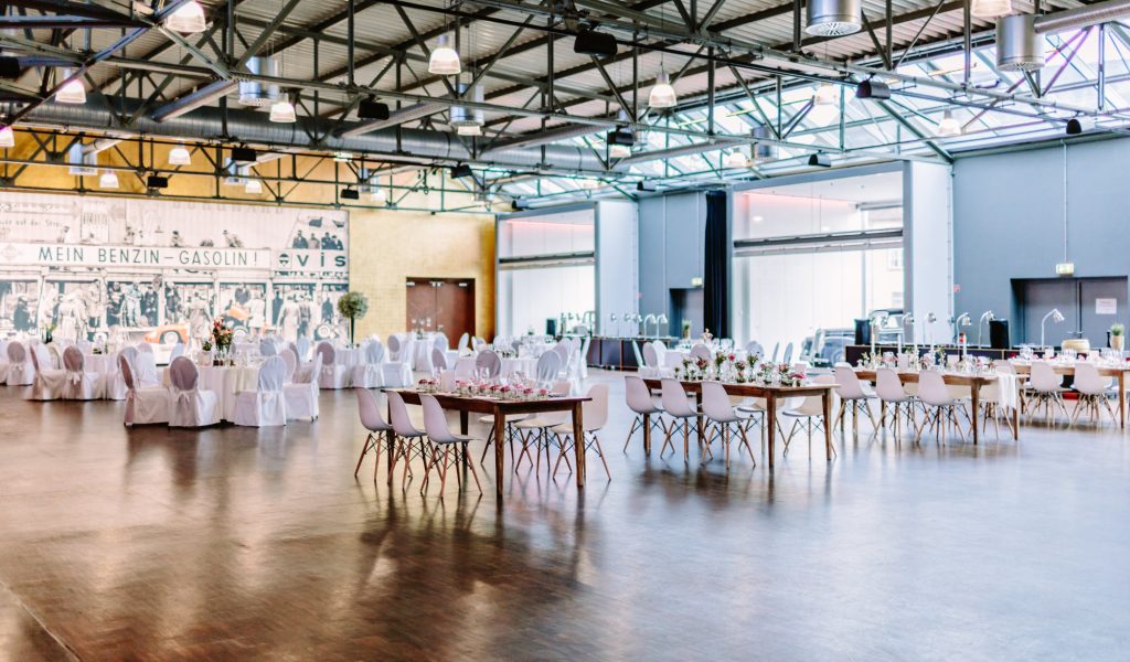 Large industrial hall with visible steel girder ceiling and pendant lights, round and long banquet tables arranged on a polished wooden floor with white chair covers and modern white chairs as well as flower arrangements in front of a wall with a historical advertising motif "MEIN BENZIN - GASOLIN!".