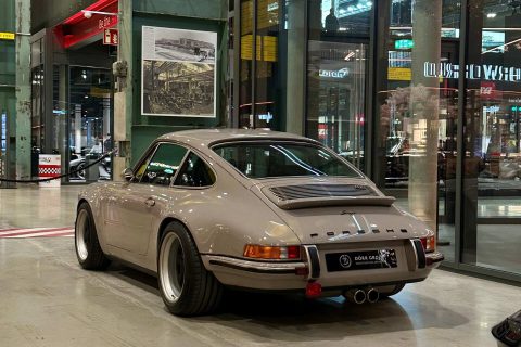 Hellbeiger klassischer Porsche 911 mit breiten Kotflügeln, parkend in einer Halle mit orangegelben Stahlträgern und Glaswänden, im Hintergrund historische Fotos an einer Säule.