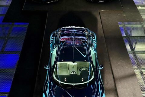 Drei Bugatti-Supersportwagen in einer Ausstellung, von oben fotografiert, auf schwarzem Teppich mit blau beleuchtetem Boden links und rechts, zwei im Hintergrund (einer silbern, einer schwarz) und ein auffällig blauer im Vordergrund mit weißem Innenraum