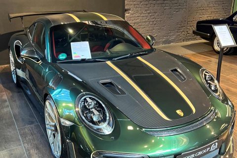Grüner Porsche 911 GT2 RS mit goldenen Rennstreifen und Karbon-Elementen in einem Showroom mit Holzboden und Backsteinwand.