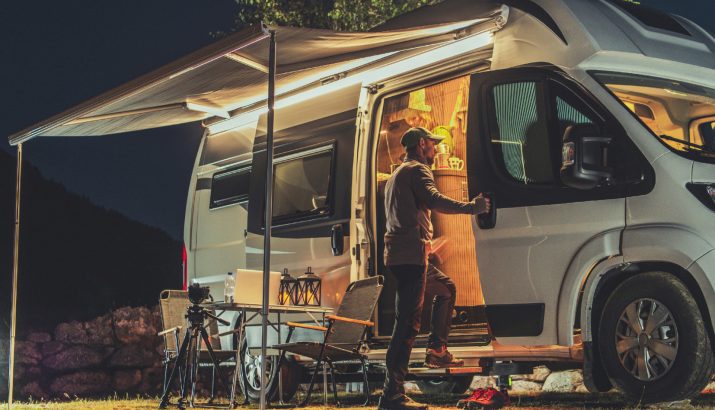 Mann öffnet die Tür eines beleuchteten Campervans bei Nacht, daneben ein Tisch mit Kamera auf Stativ, Wasserflasche und zwei Laternen, zwei Stühle unter einem ausgeklappten Markisendach auf Wiese mit steinigem Hintergrund.