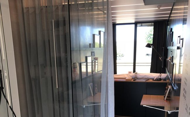 Moderner Innenraum mit schwarzer Möbelwand, Klapptisch, Stehlampe und großen Fenstern mit Blick nach draußen, teilweise durch einen transparenten Vorhang verdeckt