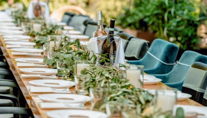 Langer, im Freien gedeckter Holztisch für viele Gäste mit weißen Tellern, Gläsern, Kerzengläsern, einer grünen Zweiggirlande als Tischläufer und Weinflaschen in einem Eisbehälter, gesäumt von blauen Samtstühlen.
