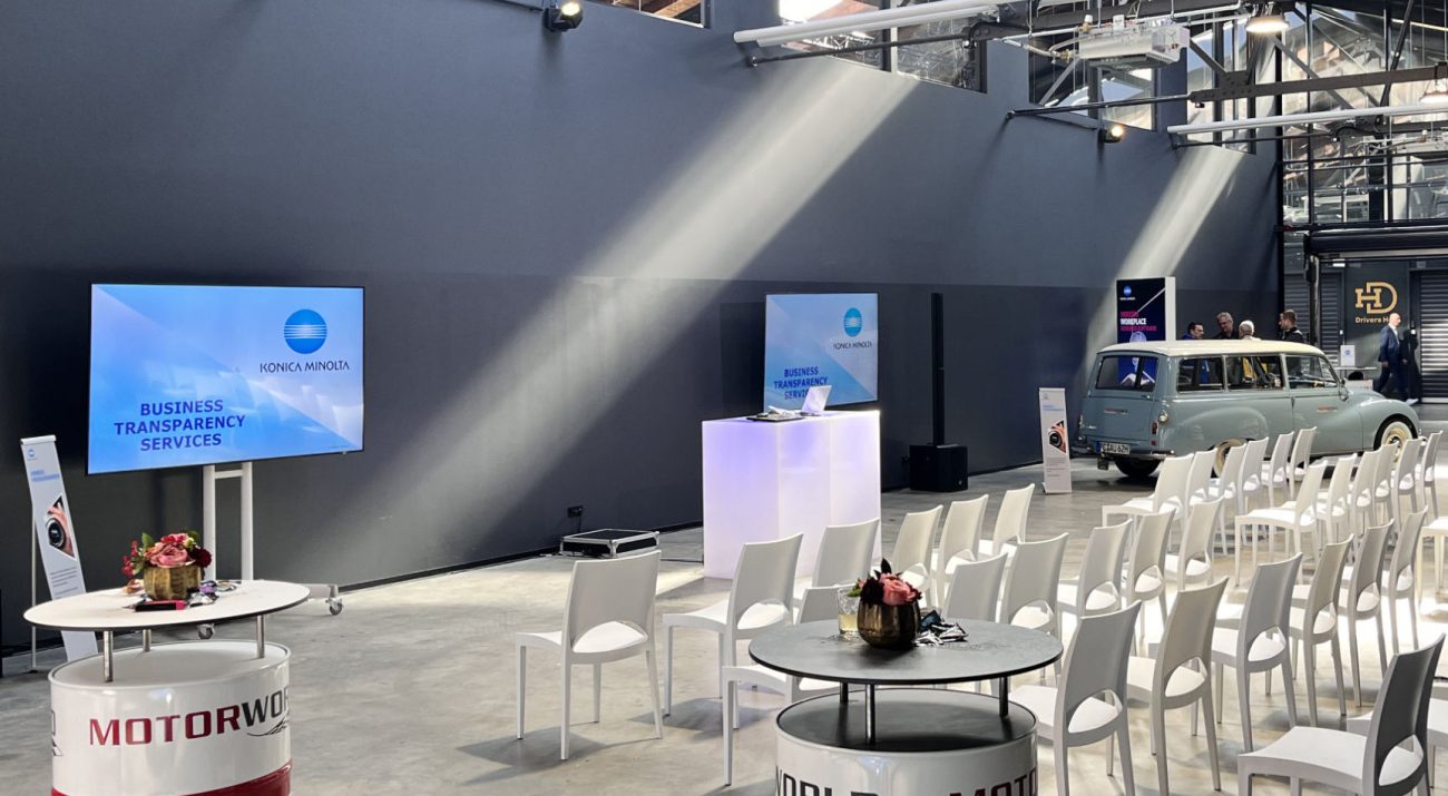 Large, bright event room with several rows of white chairs, tables made from painted oil drums with flower arrangements, two monitors with Konica Minolta Business Transparency Services and a light blue vintage station wagon on the right-hand side of the picture under visible steel beams and skylights.
