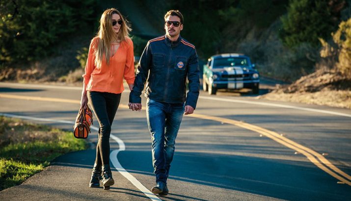 A couple is walking hand in hand on a winding road, the woman is wearing an orange blouse and black trousers, the man a dark leather jacket and jeans, a blue vintage car with white stripes can be seen in the background.