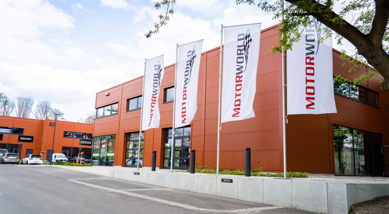 Front view of a modern, orange office building with large windows and four tall flagpoles with white flags reading MOT0RWORLD in a parking lot with several parked vehicles and a tree on the right of the picture.