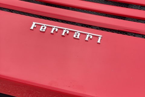 Nahaufnahme der hinteren Motorhaube eines roten Ferrari mit silbernem Ferrari-Schriftzug und dem Emblem eines springenden Pferdes auf dem schwarzen Lüftungsgitter.