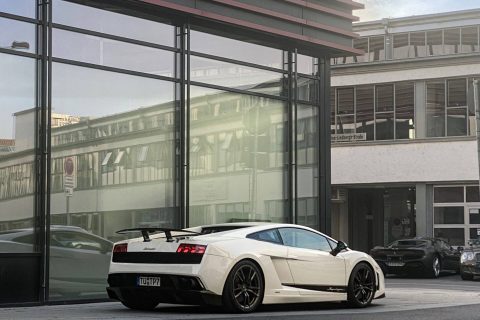 Weißer Lamborghini Gallardo mit schwarzem Heckspoiler, geparkt vor einem modernen Gebäude mit großen Glasfenstern und reflektierender Fassade.