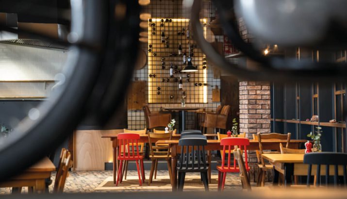 Cozy interior of a restaurant with several wooden tables and colorful chairs (red and black), an illuminated wine bottle wall at the back, a brick wall and warm hanging lamps.