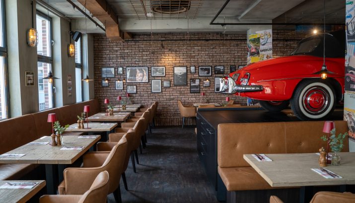 Vintage-Stil Restaurant mit braunen Ledersesseln, Holztischen, Ziegelwand mit Bildern und einem roten Oldtimer auf einer Bühne.