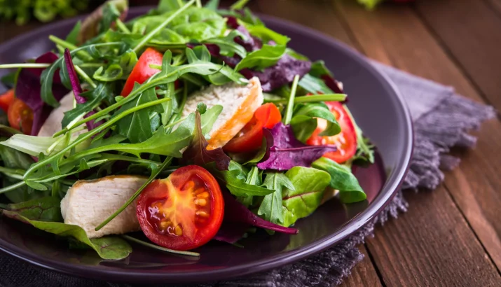 Fresh salad with chicken, tomatoes and mixed greens (arugula, mesclun, mache) on wooden background close up. Healthy food.
