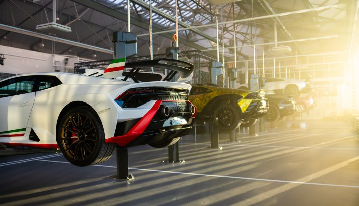 Mehrere Sportwagen auf Hebebühnen in heller Werkstatt; vorn ein weißer Wagen mit großem Heckspoiler und grün-weiß-rotem Streifen.